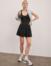black mini dress in our stretch woven fabric with stitch down front pleats, a scoop front neckline, open back detail, bodice in performance stretch fabric, and skirt side vents for easy pocket access.